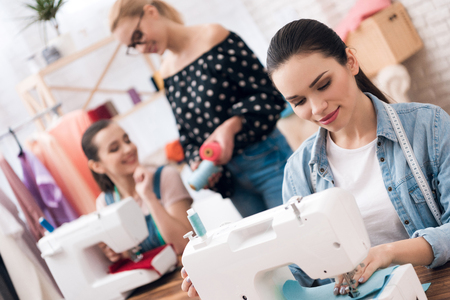 Three girls at garment factory. They are happy and fashionable. They are sitting behind sewing machines and choosing threads for new dress.の写真素材