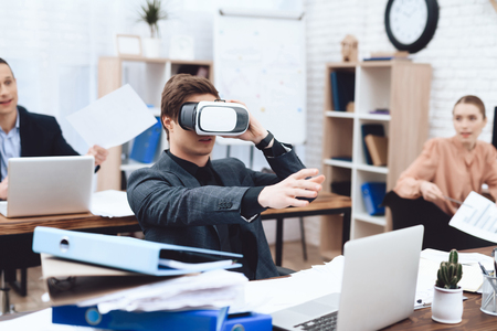 The guy has fun with virtual reality glasses. He works in a business office. His colleagues are...
