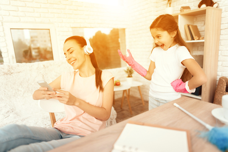 A woman is sitting at a table near a laptop and resting her legs on the table. Her daughter rearranges the house behind her. The girl is unhappy that her mother is resting and does not help her.の写真素材