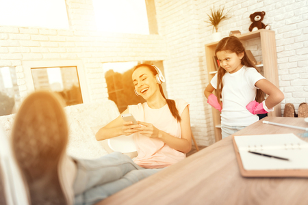 A woman is sitting at a table near a laptop and resting her legs on the table. Her daughter rearranges the house behind her. The girl is unhappy that her mother is resting and does not help her.の写真素材