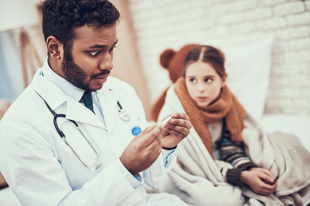 Indian doctor in white gown seeing patients at home. Doctor is taking temperature of little girl with cold.の写真素材