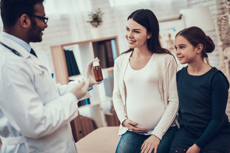 Indian doctor in white gown seeing patients in office. Doctor is giving syrup to pregnant woman with daughter.の写真素材