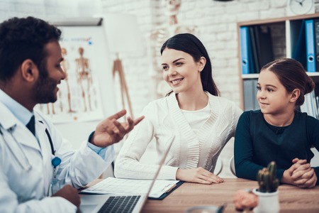 Indian doctor in white gown seeing patients in office. Doctor is talking to mother and daughter using laptop.の写真素材