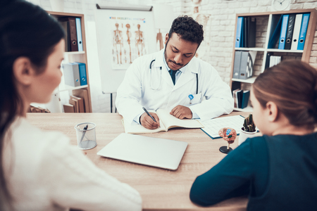 Indian doctor in white gown seeing patients in office. Doctor is taking notes listening to mother and daughter.の写真素材