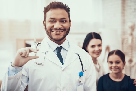 Indian doctor in white gown seeing patients in office. Doctor is posing with syringe with mother and daughter in background.の写真素材