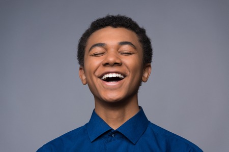 Laughing African American teenager in denim shirt. Isolated on a gray background. Transitional age...