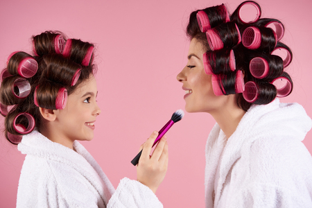 Teen girl with curlers doing makeup to mom. Isolated on pink background. Studio portrait.の写真素材