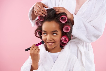 Beautiful mother and daughter in bathrobes make hairstyles with hair curlers. Little girl doing make up. Isolated on pink background.の写真素材