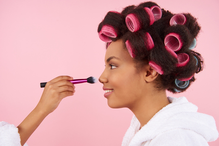 African American little girl doing makeup with mom with hair curlers. Isolated on pink background.の写真素材
