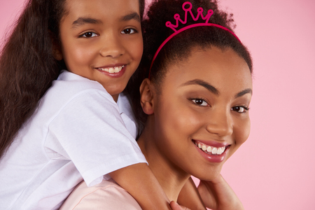 Afro American daughter on piggy back ride with mother. Isolated on pink background. Studio portrait.の写真素材