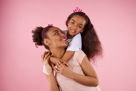 Afro American daughter on piggy back ride with mother. Isolated on pink background. Studio portrait.の写真素材