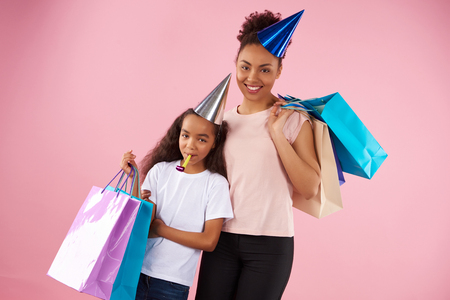 Afro American mother and daughter in holiday caps keep paper glossy bags. Shopping and consumerism concept. Isolated on pink background. Studio portrait.の写真素材
