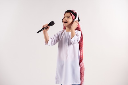 Arabian boy in keffiyeh with microphone sings karaoke. Isolated on white background. Studio portrait.の写真素材