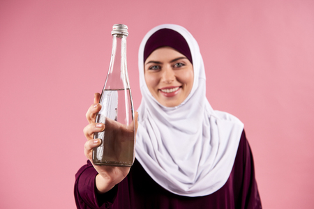 Arab woman in hijab holds bottle of water. Clean water concept. Isolated on pink background.の写真素材