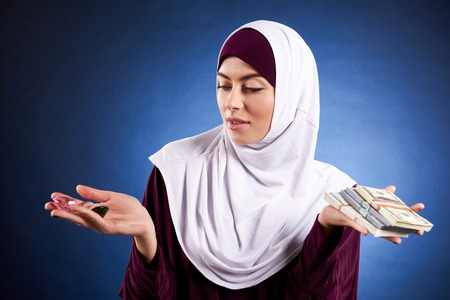 Arab woman in hijab is holding bundle of dollars and precious stones. Isolated on blue background.の写真素材