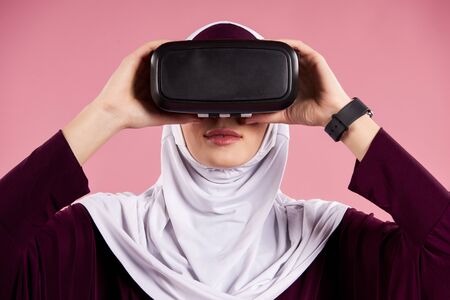 Arab woman puts on virtual reality glasses. Future technology concept. Isolated on pink background.の写真素材