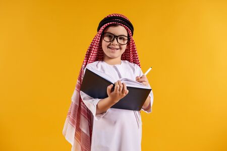 Arab happy boy with glasses makes notes in notebook. Children education concept. Isolated on yellow background.の写真素材