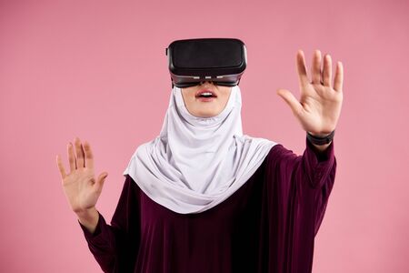Arab woman in hijab interacts with glasses of virtual reality. Future technology concept. Isolated on pink background.の写真素材