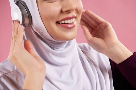 Close up. Arab woman in hijab listens to music on headphones. Isolated on pink background.の写真素材
