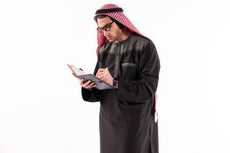 Arab man in keffiyeh makes notes in notebook. Isolated on white background. Studio portrait.の写真素材