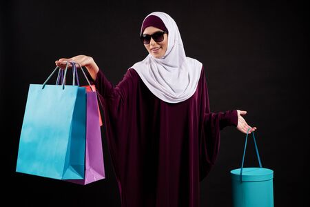 Arab woman in hijab and sunglasses holds paper colored bags. Shopping and consumerism concept. Isolated on black background.の写真素材