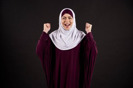 Arab woman in hijab yells with clenched fists. Isolated on black background. Studio portrait.の写真素材