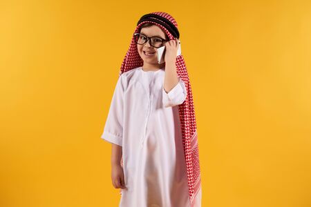 Arab smart boy in kufiya talking on smartphone. Isolated on yellow background. Studio portrait.の写真素材