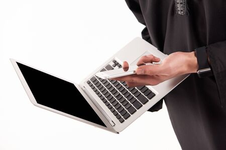 Close up. Arab confident businessman in keffiyeh holds laptop with smartphone. Isolated on white background.の写真素材