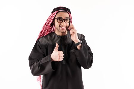 Happy Arab businessman in keffiyeh talks on cellphone and showing thumbs up. Cellular technology concept. Isolated on white background.の写真素材