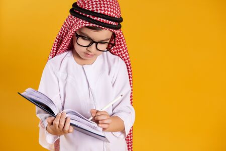 Arab happy boy with glasses makes notes in notebook. Children education concept. Isolated on yellow background.の写真素材