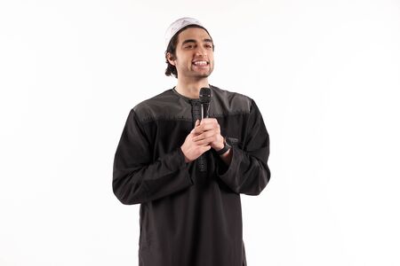 Arab man in ethnic attire stands with microphone. Isolated on white background. Studio portrait.の写真素材