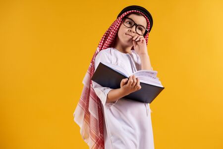 Arab happy boy with glasses makes notes in notebook. Children education concept. Isolated on yellow background.の写真素材