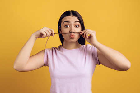 Young girl puts strand of hair under nose, showing false mustache. Isolated on yellow background. Studio portrait.の写真素材