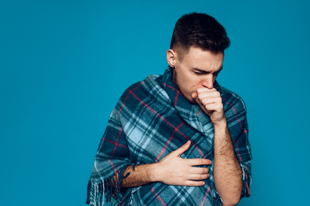 Young ailing guy with influenza virus coughs isolated on blue background. Cold. Treatment of bronchitis. Studio portrait. Flu.の写真素材
