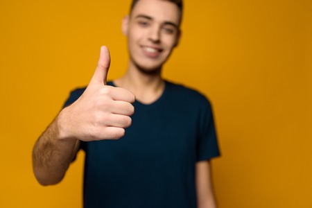 Young handsome thin guy shows thumbs up isolated on yellow background. Studio portrait.