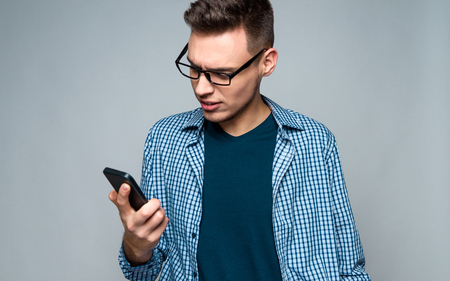 Young smart guy with glasses is looking at smartphone screen. isolated on grey background. Studio portrait.の写真素材