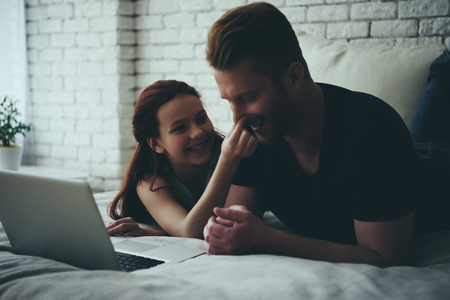 Single father and daughter lie on bed and look at screen of laptop. Family leisure concept. Fatherhood.の写真素材