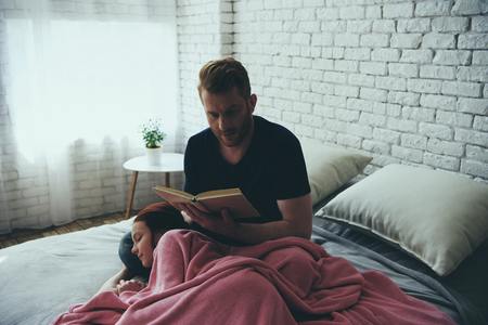 Redheaded single father reads book while daughter sleeps on his lap on bed. Family leisure concept. Fatherhood.の写真素材