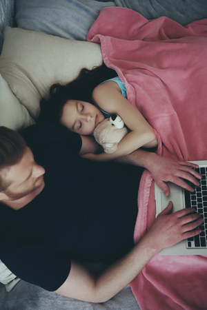 Top view. Red haired single father works with laptop, lying next to sleeping daughter. Time to sleep. Fatherhood.の写真素材