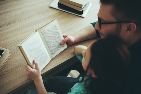 Red haired father reads book with teenage daughter sitting at table. Child education concept. Fatherhood.の写真素材