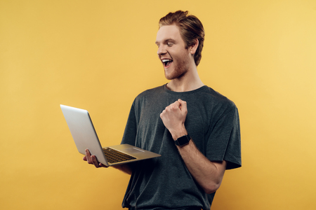Happy Guy Celebrating Success Using Laptop. Smiling Bearded Modern Caucasian Man Holding laptop and Showing Fist. Isolated on Orange Background. Concept of Humans Emotionsの写真素材