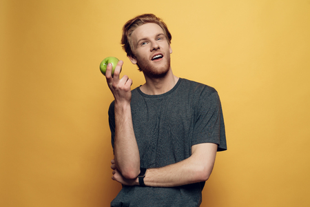 Happy Joyful Man Holding Green Apple and Thumbs Up. Handsome Man Looking at Camera with Amazed Expression Showing Thumbs up. Isolated on Orange Background. Concept of Human Emotionsの写真素材