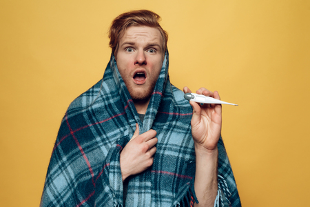 Guy Wrapped in Plaid Pointing on Thermometer. Sick Guy in Checkered Blue Wrap looks at camera. Ill Handsome Person Feels Uncomfortably Unhealthy Isolated on Yellow Background. Concept of Coldの写真素材