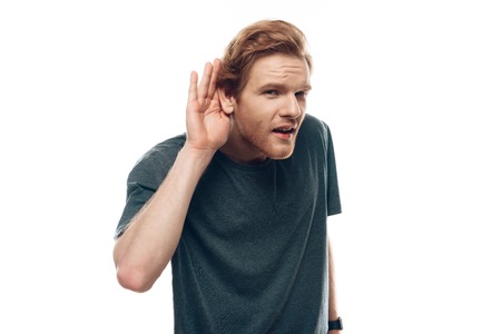 Portrait of Young Man with Hearing Problem. Ginger Serious Young Guy in Casual Clothes has Questionable Face Expression Holding Hand Near Ear Isolated on White background. Concept of Human Emotionsの写真素材