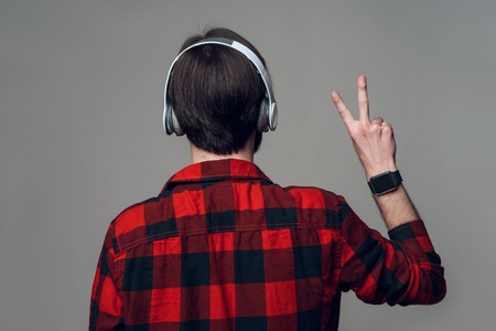 Back View. Man Listening to Music with Headphones. Young Brunette Guy in Red Checkered Shirt shows gesture victory sign. Isolated on Gray Background. Concept of People Emotionsの写真素材