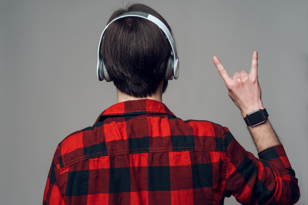 Back View. Man Listening to Music with Headphones. Young Brunette Guy in Red Checkered Shirt shows gesture rock sign. Isolated on Gray Background. Concept of People Emotionsの写真素材