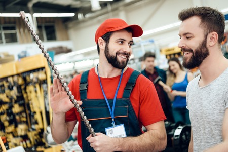 Salesman in red shirt and baseball cap is showing bearded client new giant drill in power tools store.の写真素材