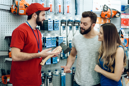 Salesman in red shirt and baseball cap is showing couple of clients new axe in power tools store.の写真素材