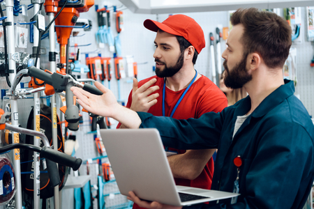 Two salesmen in red shirt and baseball cap and blue robe are checking tooks inventory with laptop in power tools store.の写真素材