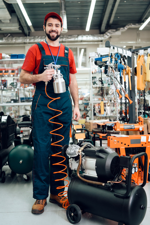 Salesman in red shirt and baseball cap is posing with compressor paint sprayer on foreground in power tools store.の写真素材